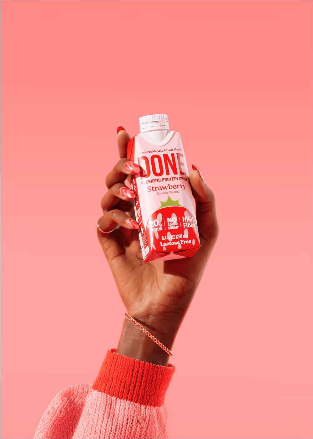 Hand holding a DONE Drinks Strawberry protein shake with red-pink nails and a pink sweater, against a vibrant pink background.