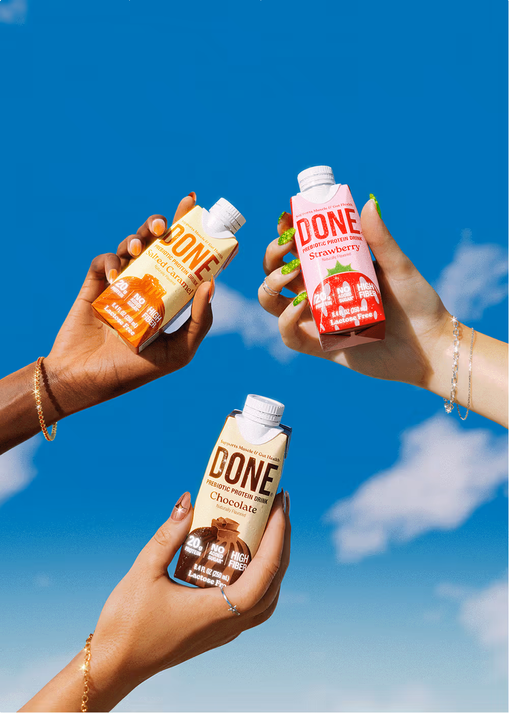 Three hands holding DONE Drinks protein beverages in Salted Caramel, Strawberry, and Chocolate flavors against a bright blue sky.