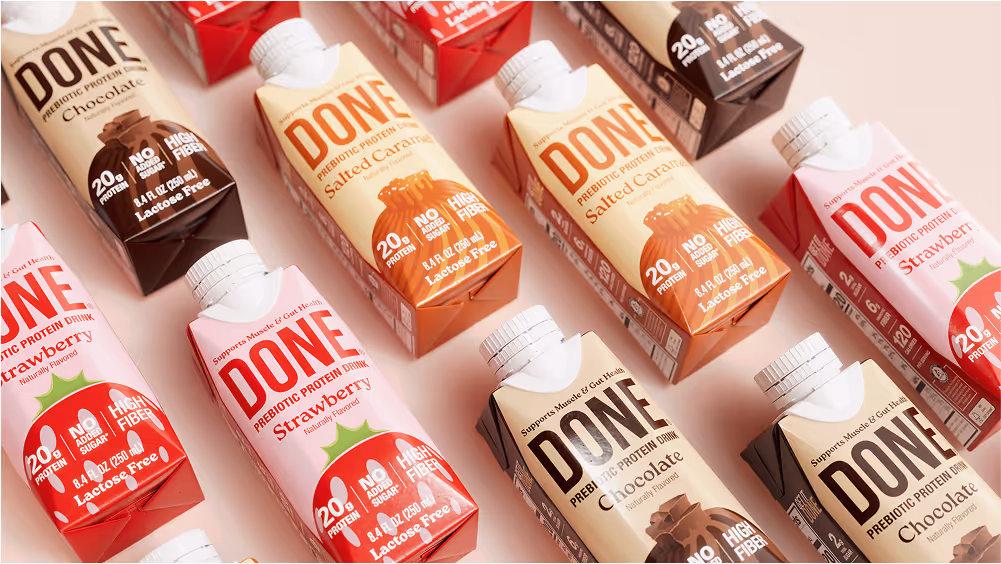 Multiple DONE Drinks prebiotic protein cartons in Chocolate, Salted Caramel, and Strawberry flavors arranged in a grid on a light pink background.