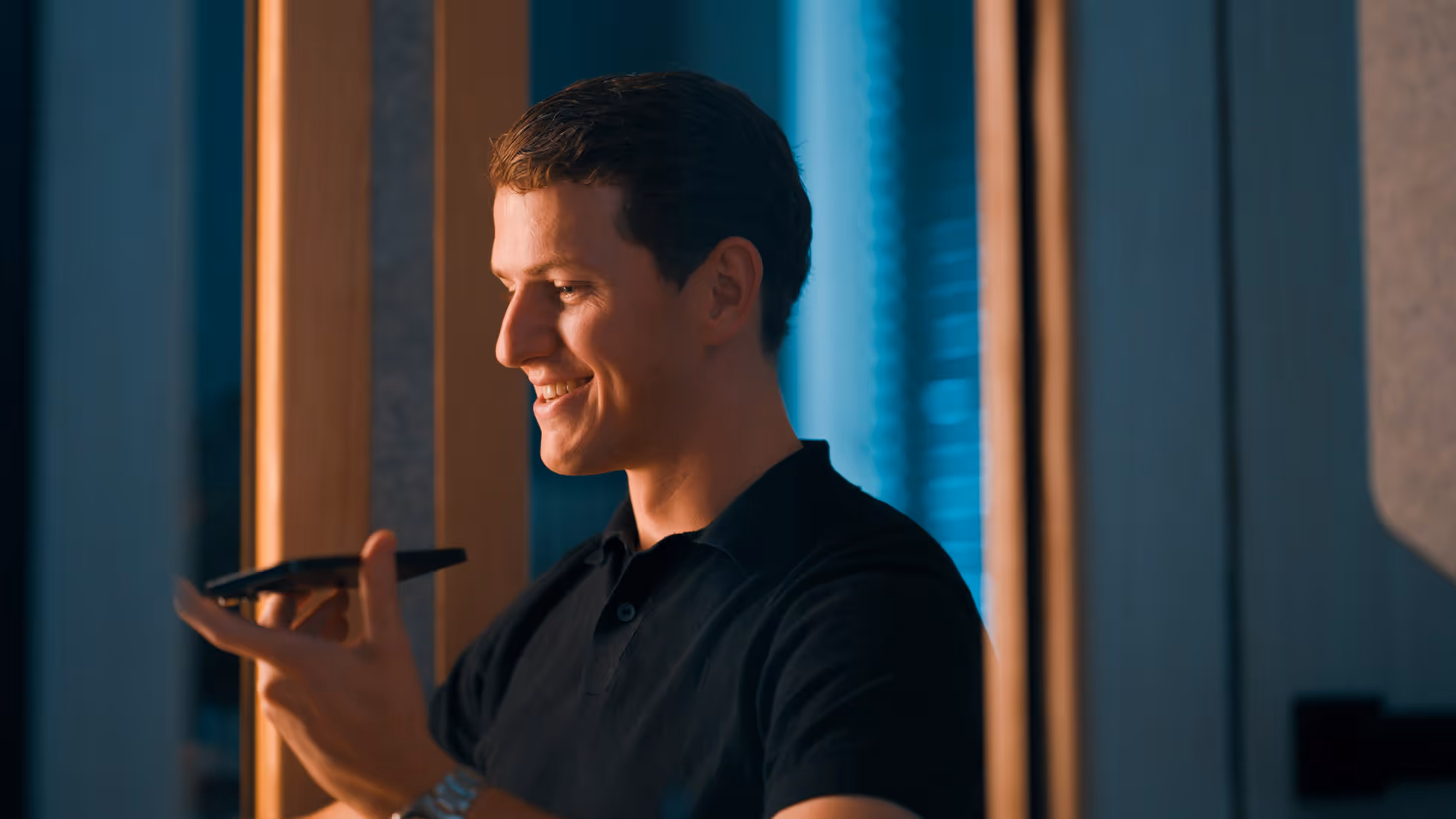Smiling man in a black shirt holding a smartphone and speaking into it indoors.