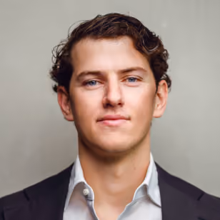 Portrait of a young man with curly brown hair wearing a dark blazer and white shirt, looking directly at the camera.