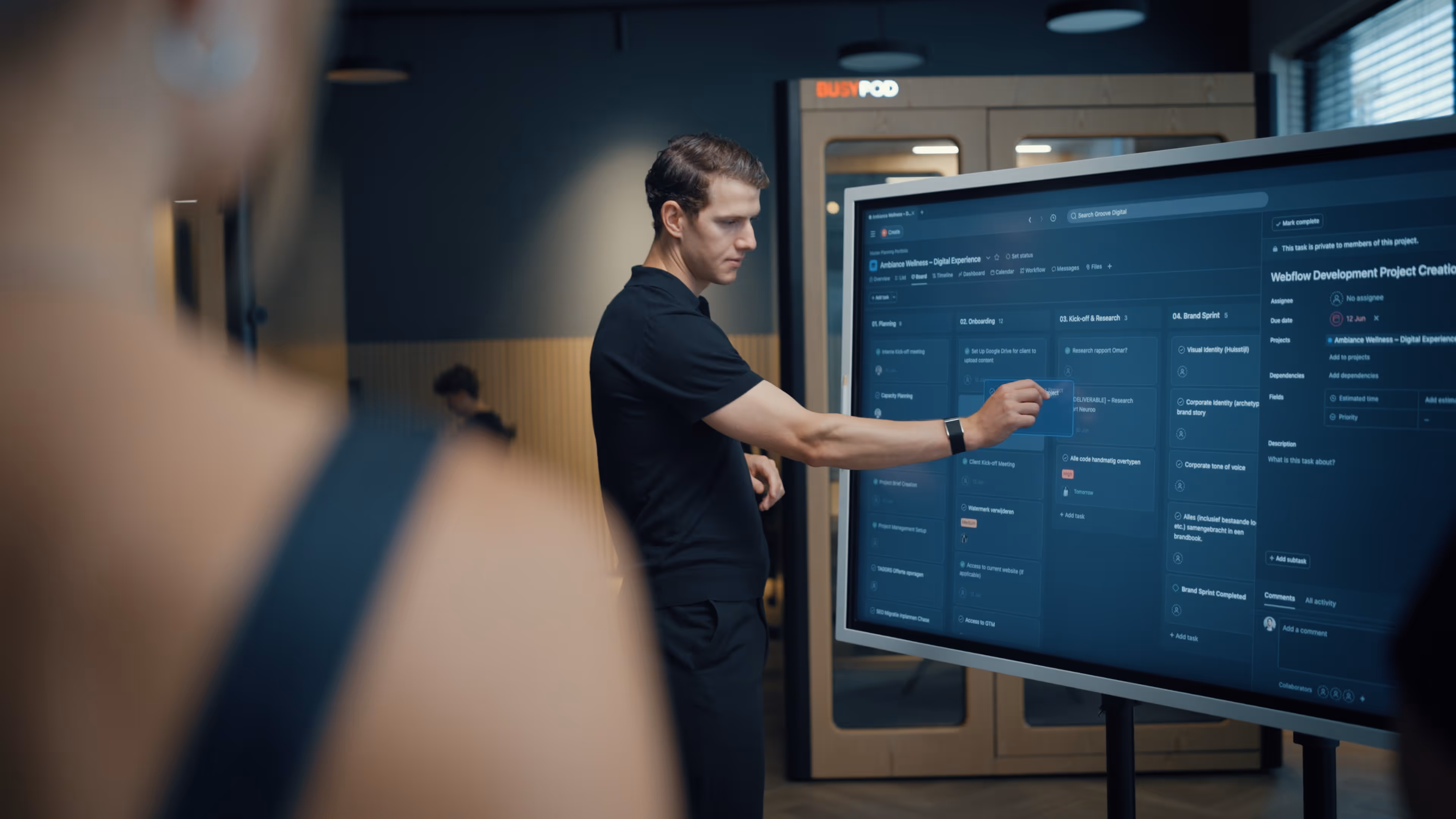 Man in black shirt interacting with a large touchscreen displaying a project management dashboard in an office setting.