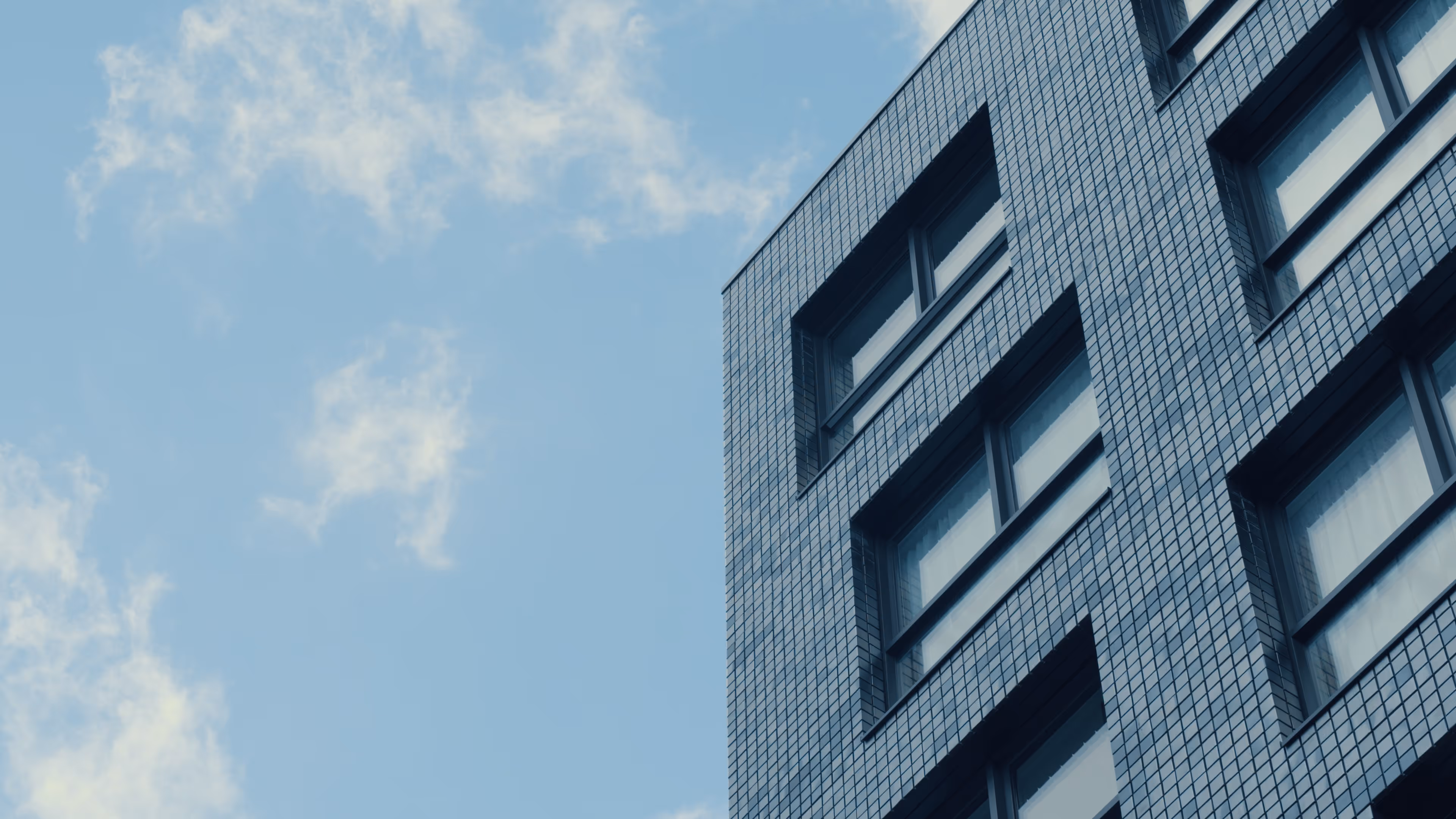 Modern building corner with blue square-tiled facade and multiple windows under a partly cloudy sky.