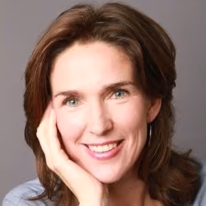 Smiling woman with shoulder-length brown hair resting her face on her hand against a plain background.