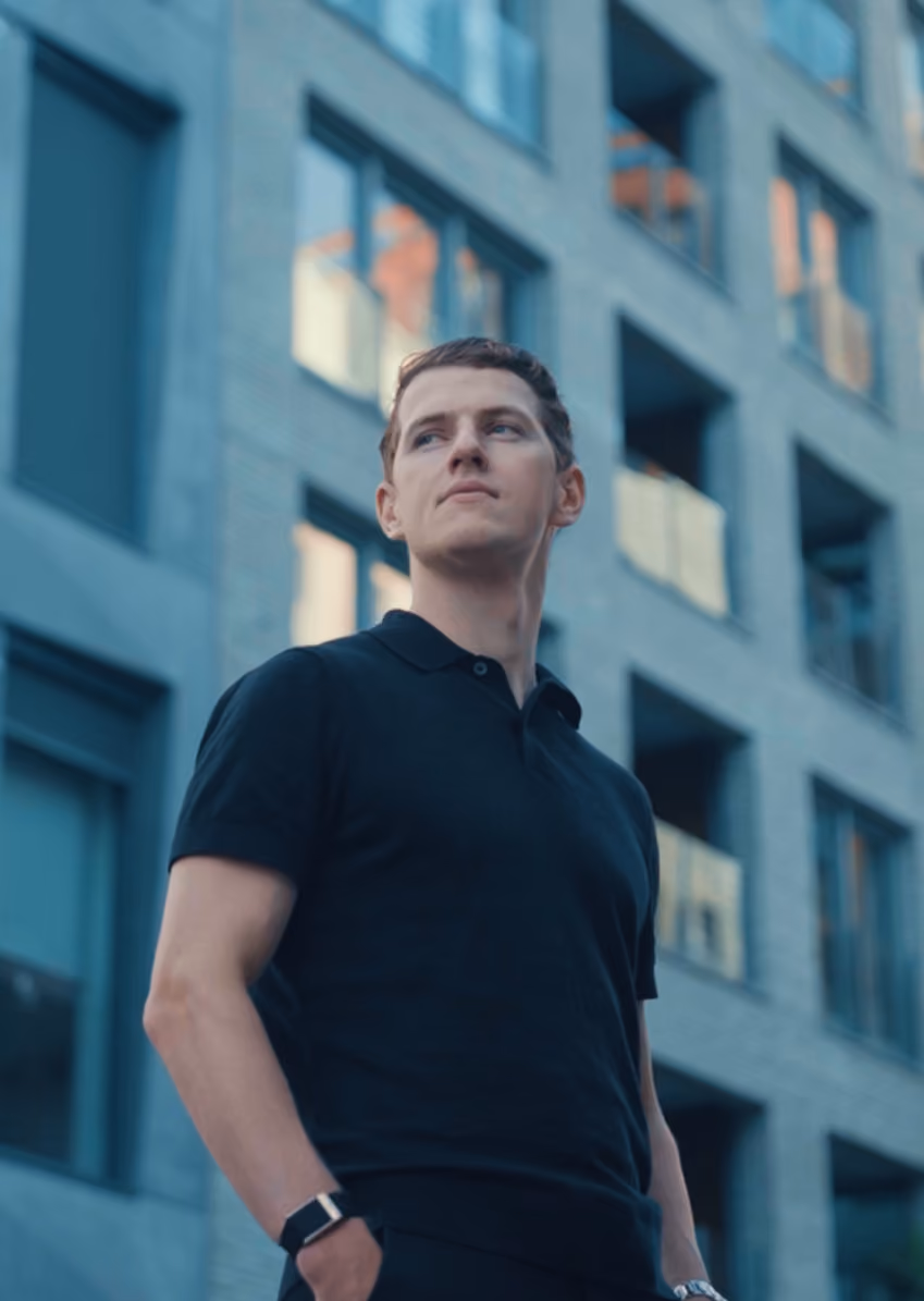 Young man in a black polo shirt standing confidently with hands in pockets in front of a modern building with large windows.