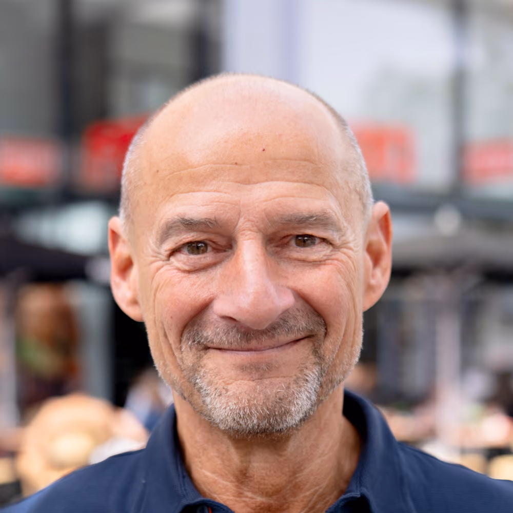 Smiling bald middle-aged man with trimmed beard wearing a navy blue shirt outside in a city setting.