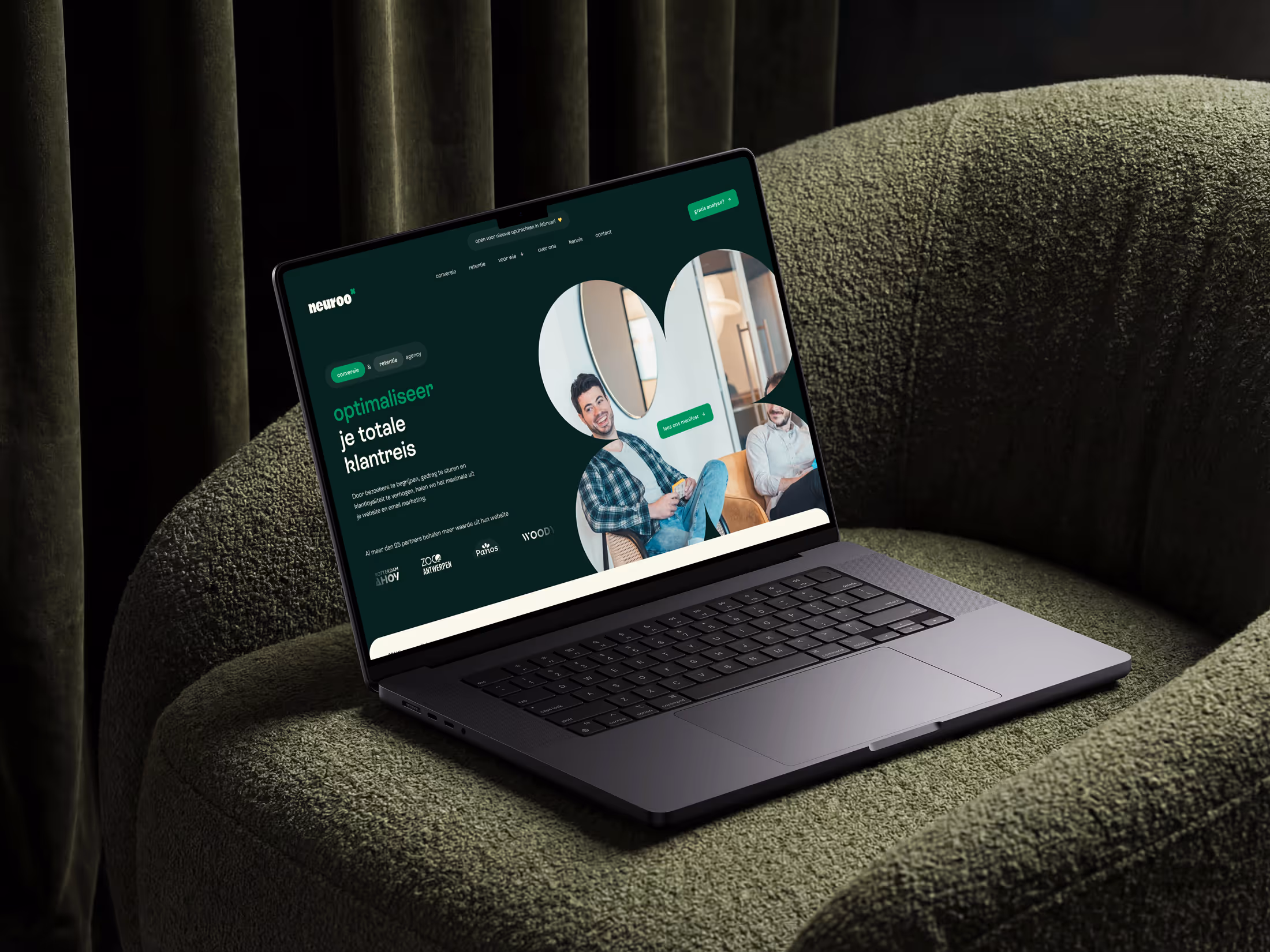 Laptop on green chair showing a website with text and two men sitting in a bright room.