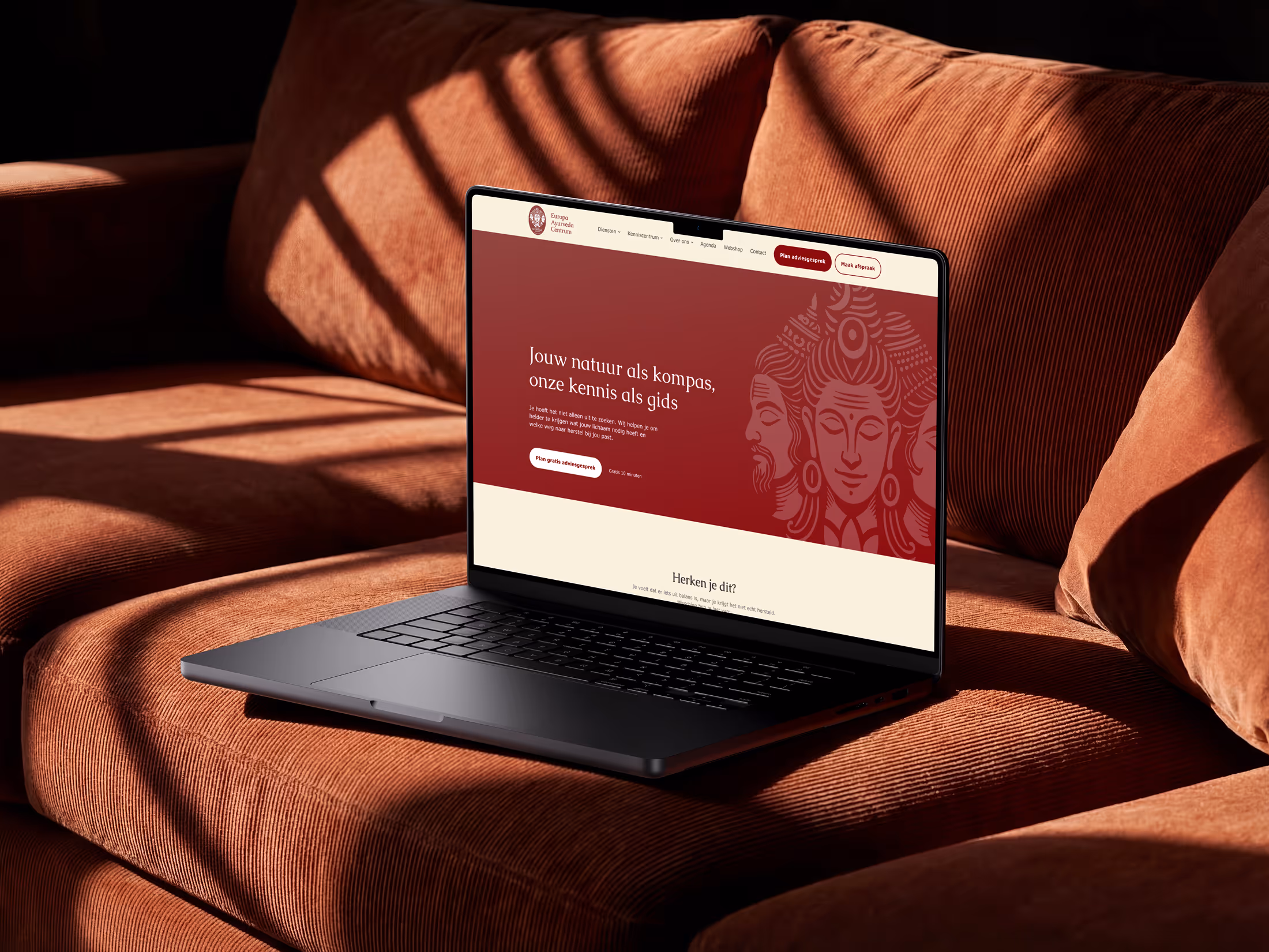 Laptop on orange velvet couch displaying a website with red and cream design and text in Dutch.