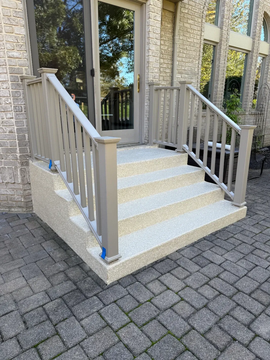 Front porch steps and small landing with a clean beige coating, white railings, and paver walkway at the base.