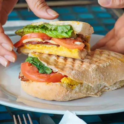 Close-up of a toasted sandwich filled with egg, tomato, lettuce, and bacon, being pulled apart by two hands over a white plate.