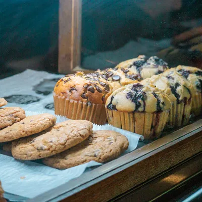 Assorted cookies and muffins displayed in a bakery case, including chocolate chip and blueberry options, on a tray lined with parchment paper.