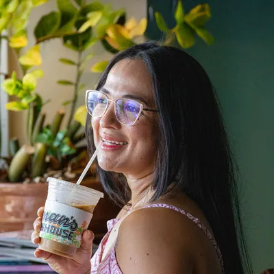 Smiling woman wearing glasses enjoying an iced coffee from Paperman’s Coffeehouse, standing near a window with lush plants in the background.