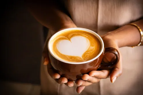 Close-up of a brown mug with heart-shaped latte art, gently held by both hands in a warm, soft light.