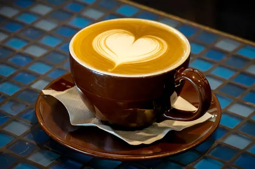 Cappuccino with heart-shaped latte art served in a brown cup and saucer, placed on a blue tiled café table with a napkin.