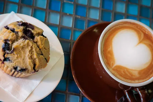 Blueberry muffin on a white plate with a napkin, next to a cappuccino with heart latte art on a tiled blue table.