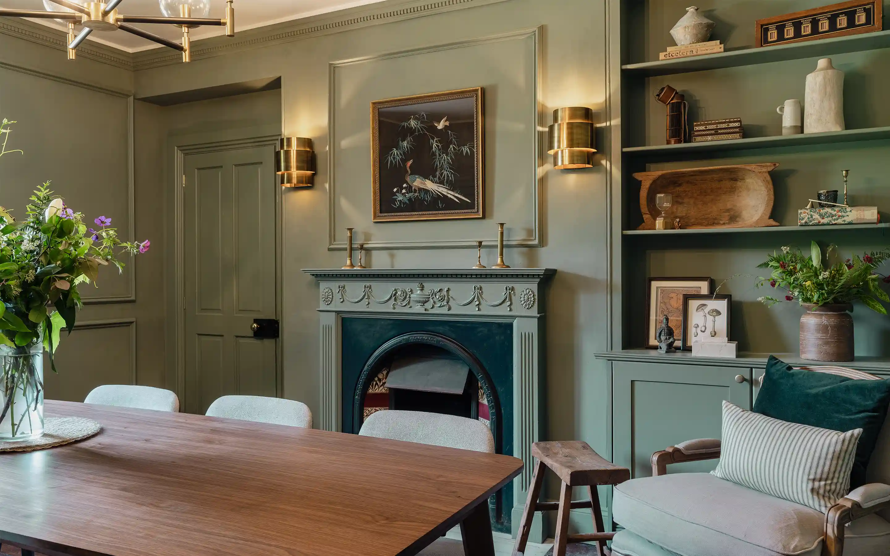 Elegant green dining room with carved fireplace, brass sconces, wooden table, and styled shelves with art and decor.