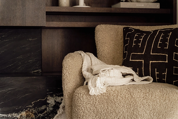 Warm modern living room with dark wood paneling, minimalist shelves, a boucle armchair, and a black marble fireplace.