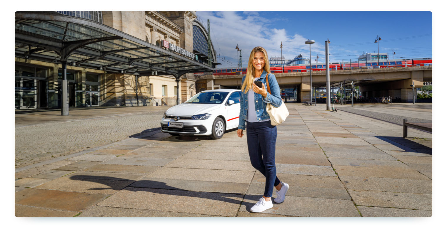 A blonde woman with a smartphone, standing in front of a Flinkster sharing car at a train station, is smiling in the camera