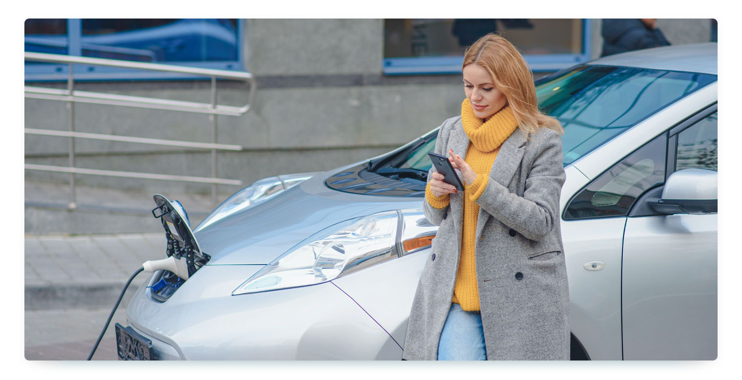 A blonde woman, wearing a yellow sweater and a grey jacket, ist standing in front of a silver car that's currently charging with her smartphone in her hands