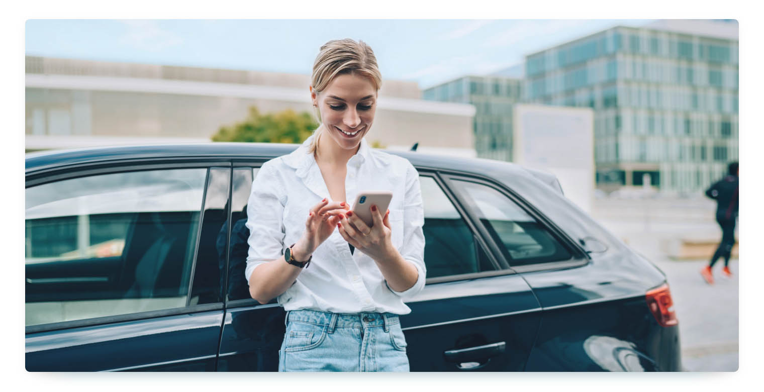 A woman leans against a dark car, smiles and types something on her smartphone