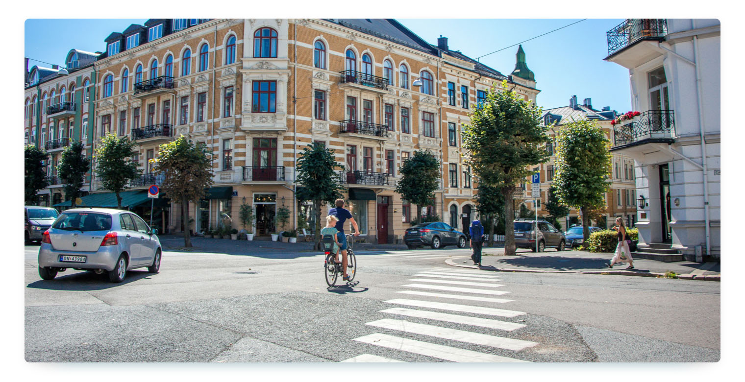 Urban street scene with classic European architecture, featuring a cyclist transporting a child, cars on the road, and pedestrians crossing at a zebra crossing.