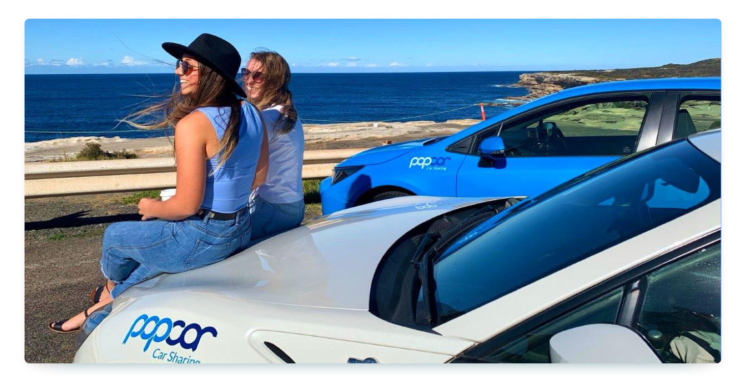 Two women are sitting on the hood of a white Popcar vehicle, with another blue Popcar car parked behind them. They are looking out over the ocean, with a cliffside visible in the background on a sunny day.