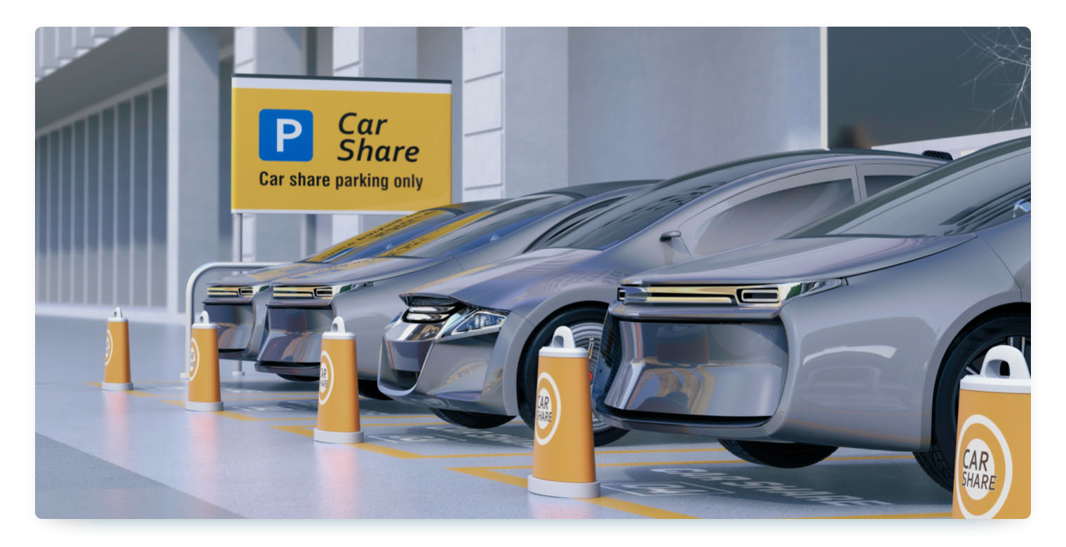 A row of modern, silver-colored electric vehicles parked in a car-sharing lot, with a prominent "Car Share" sign in the background. The parking spots are marked with yellow lines and cones labeled "CAR SHARE."
