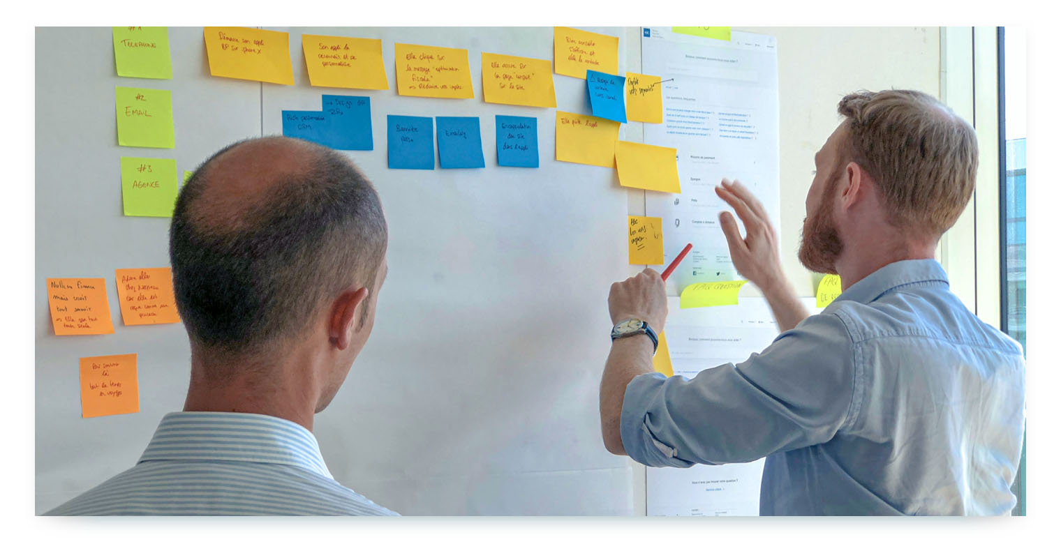 Two men in striped shirts are standing in front of a whiteboard covered with yellow and blue sticky notes. The man on the right, seen from the side, is pointing at a sticky note with a red marker. The other man, seen from the back, is looking at the board.