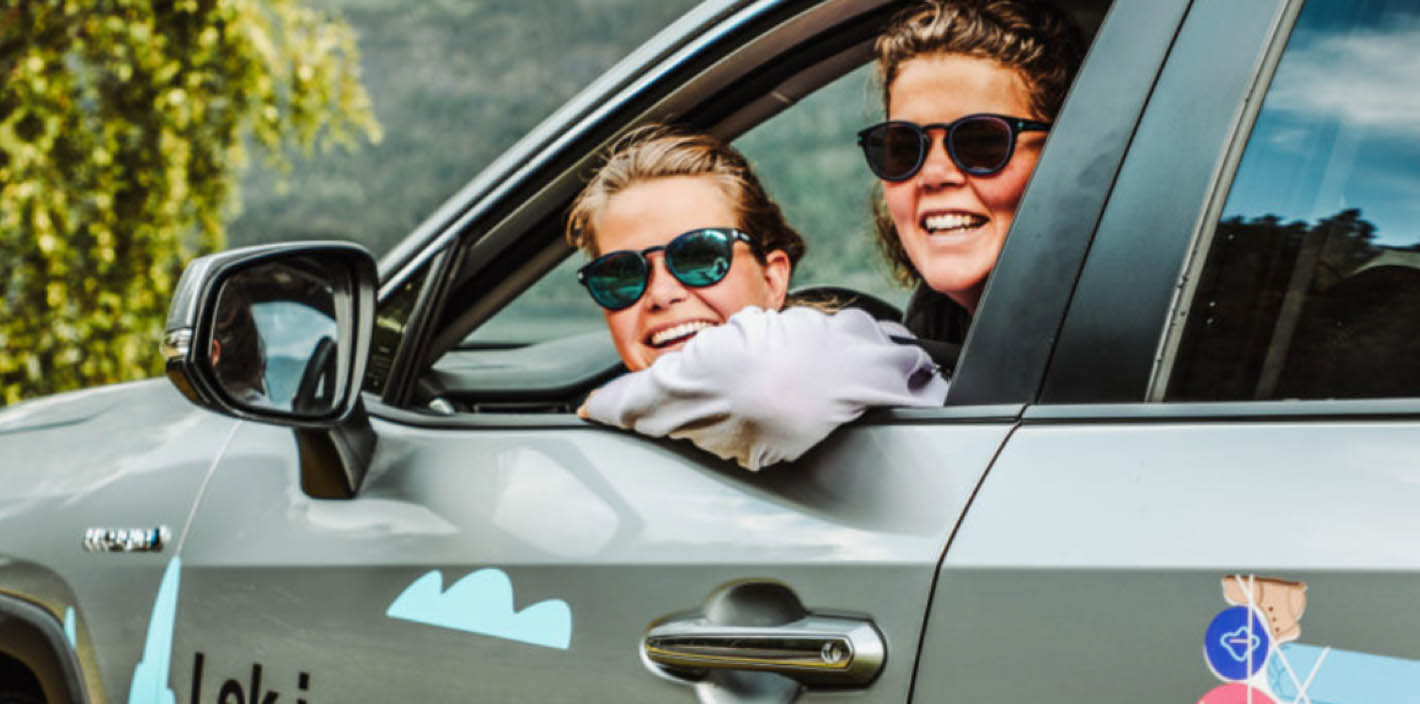 Two smiling people wearing sunglasses are leaning out the passenger side window of a gray car. The car has light blue cloud stickers and a logo on the side.