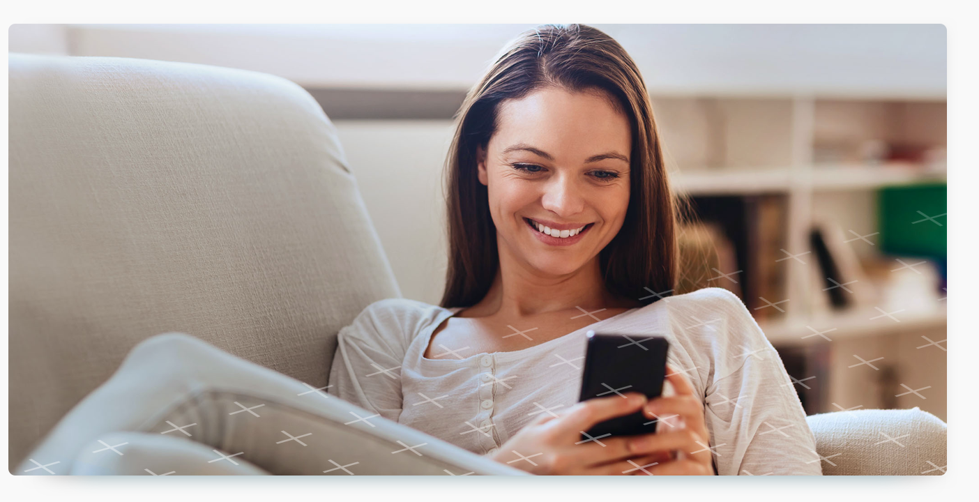 A woman with brown hair smiles while sitting on a light-colored armchair and looking at a smartphone she is holding.