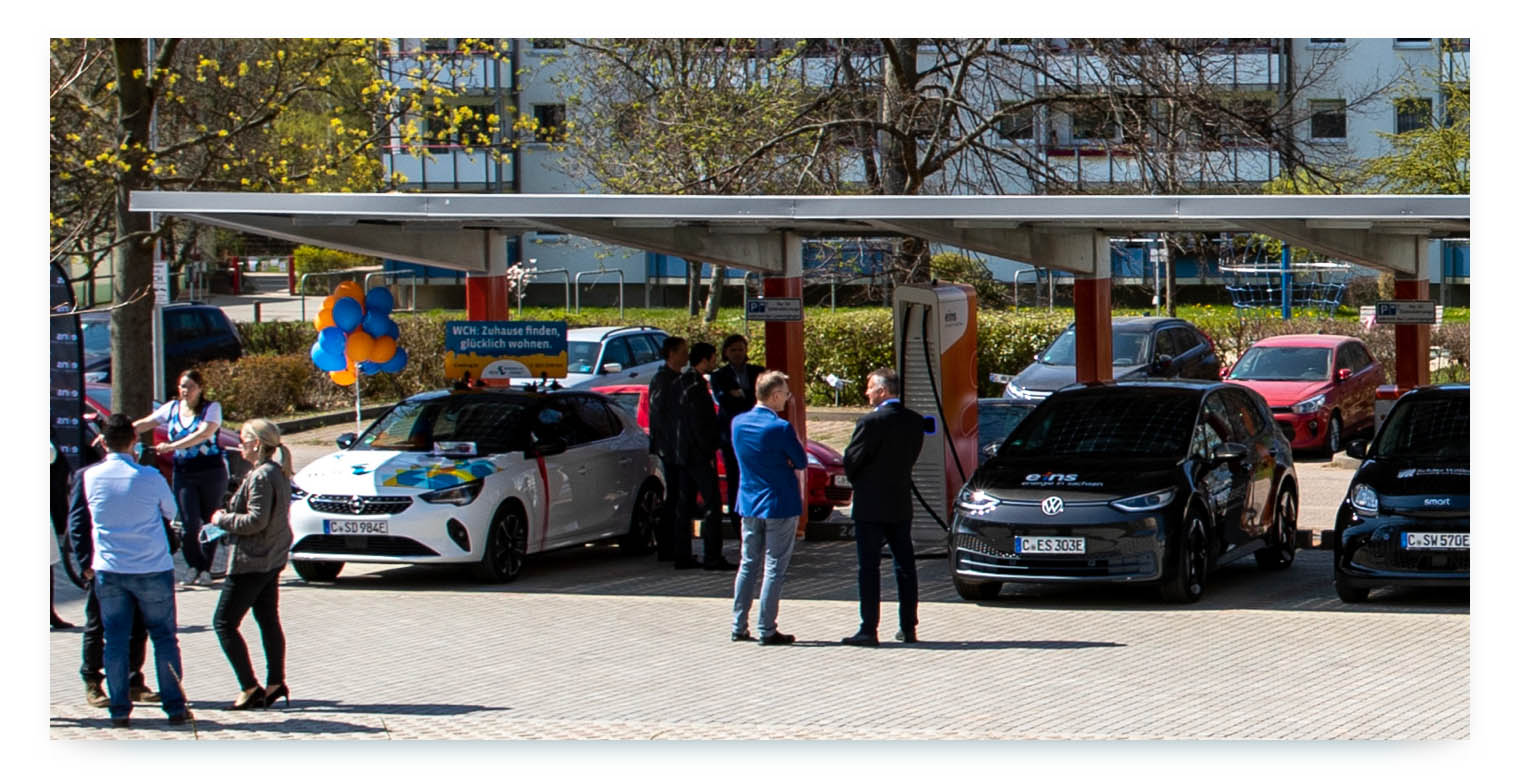 A group of people are gathered around two electric cars at a charging station. A row of balloons is tied to a sign in the background. The cars are parked in front of a building.