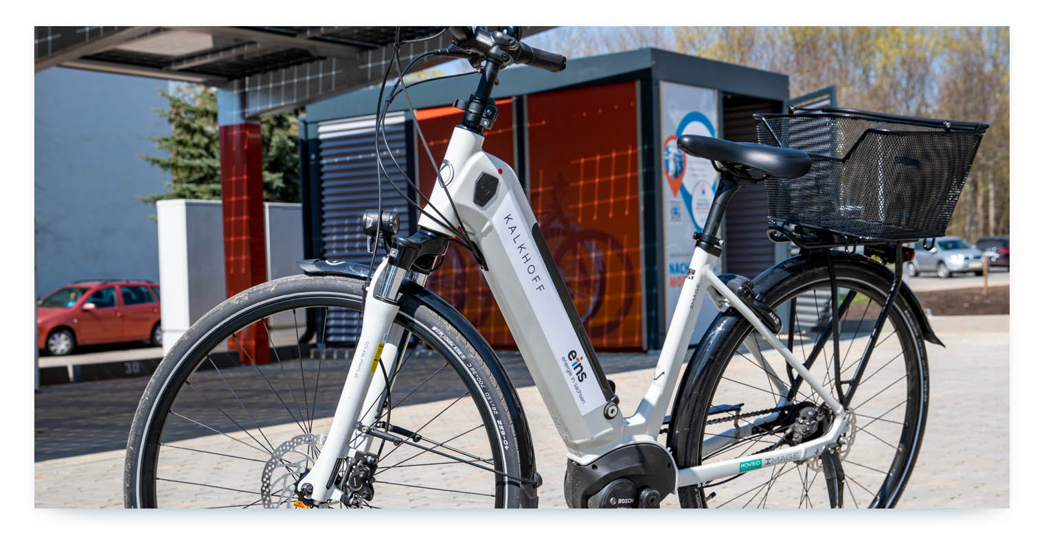 A close-up shot of an electric bicycle parked outside. The brand name KALKHOFF is visible on the frame, as well as a logo for the company "eins." A red car is visible in the background, and the bicycle has a black basket on the back.