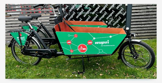 A bright green cargo bike from the "wupsi" bikesharing service. The bike's cargo box features a graphic showcasing connections to cars, buses, and trains, demonstrating a multi-modal partnership that integrates bikesharing with other forms of public and shared transport.
