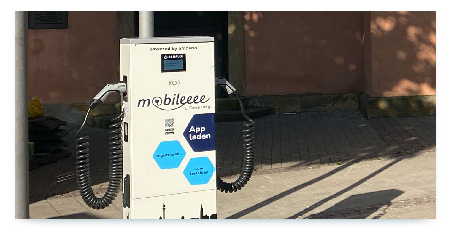 A charging station for shared electric cars with the "mobileeee E-Carsharing" logo and branding. The station also shows the "amperio" logo, indicating a partnership between a shared mobility operator and a charging infrastructure provider. This illustrates how strategic partnerships are crucial for building the necessary infrastructure for e-carsharing.