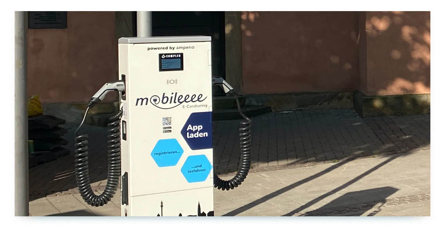 A charging station for shared electric cars with the "mobileeee E-Carsharing" logo and branding. The station also shows the "amperio" logo, indicating a partnership between a shared mobility operator and a charging infrastructure provider. This illustrates how strategic partnerships are crucial for building the necessary infrastructure for e-carsharing.