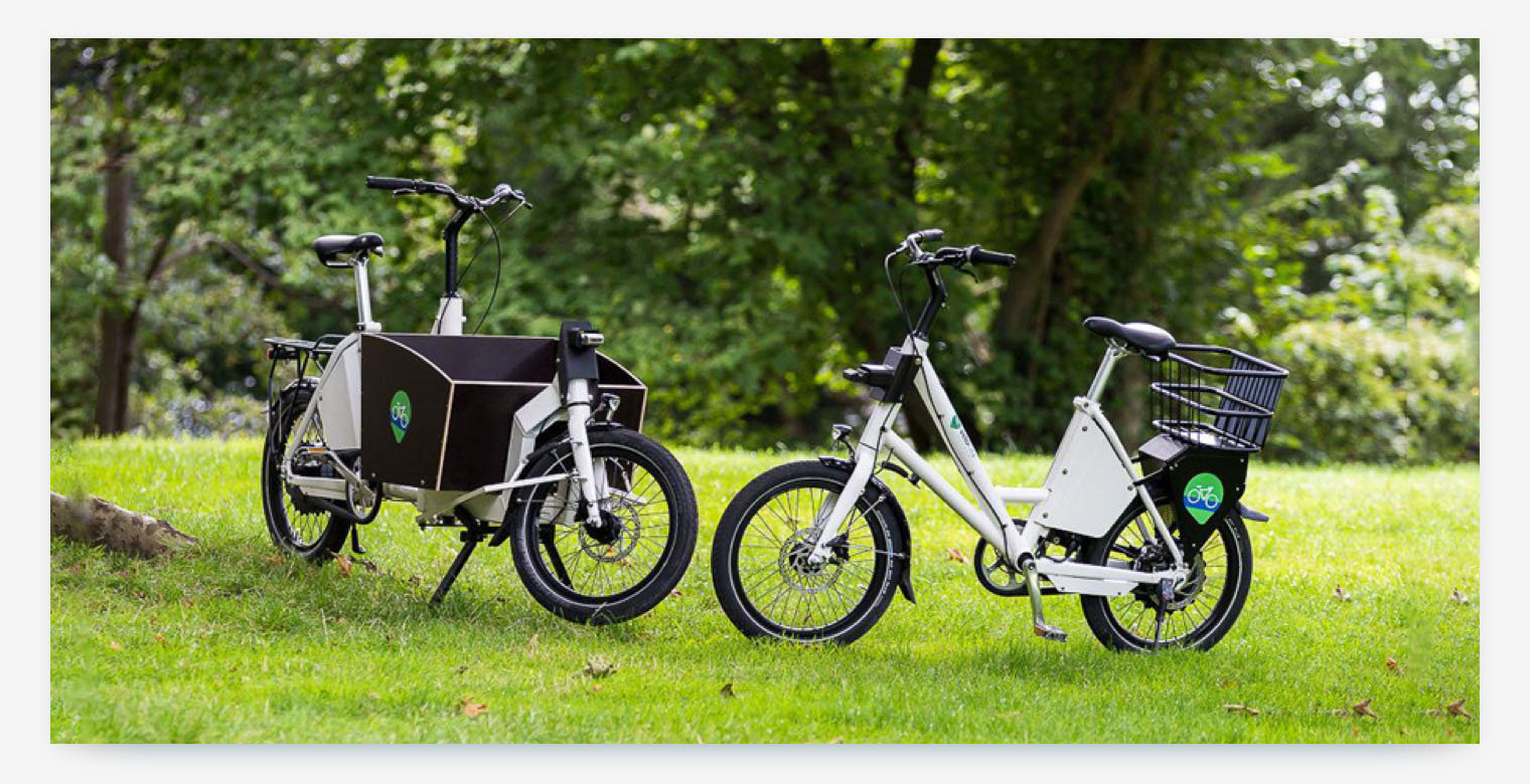 Two different types of shared mobility bikes, a cargo bike and an e-bike, are parked side-by-side in a park. 