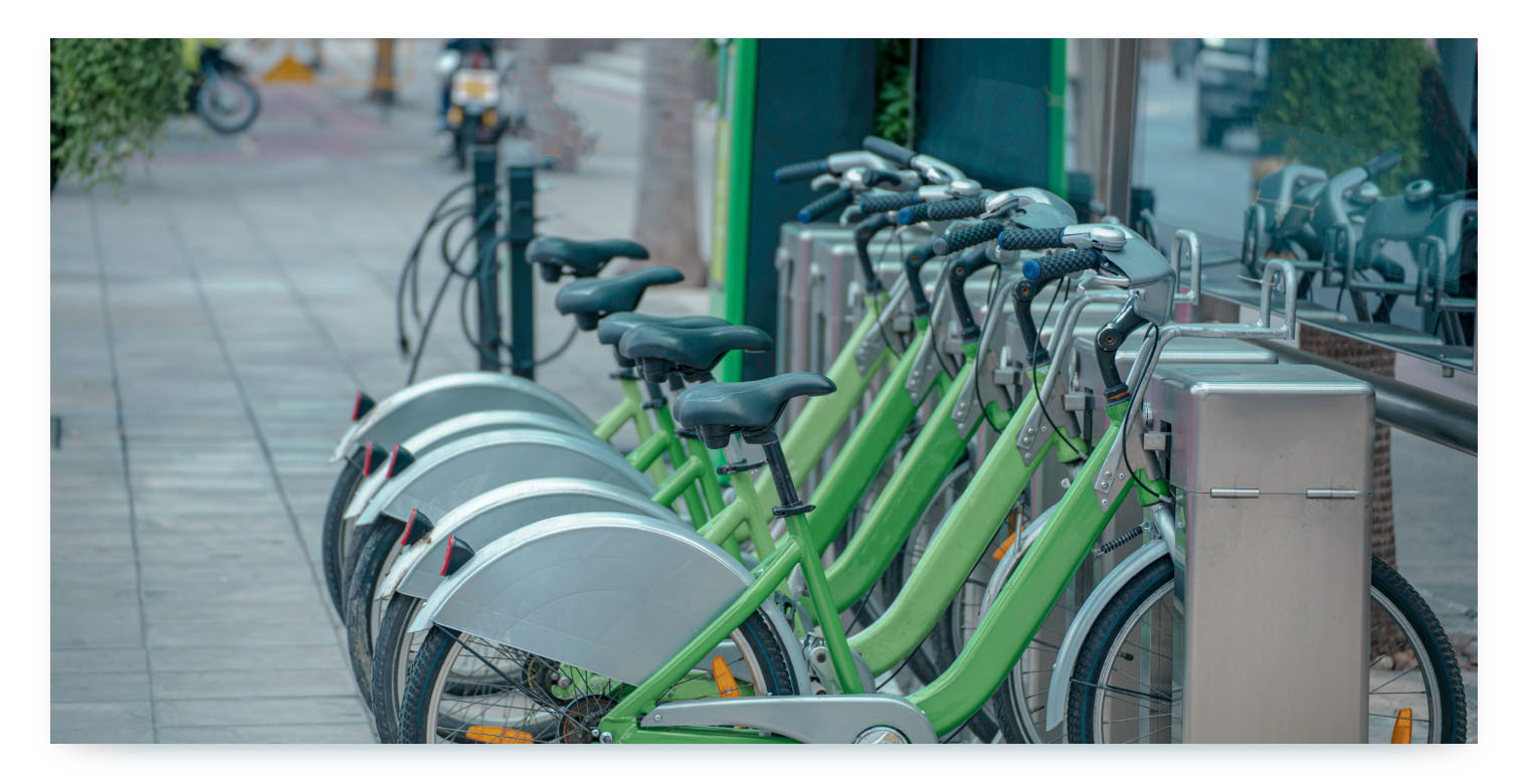 A row of green and silver shared bicycles are neatly parked and locked at a bike rental station on a city sidewalk. The sharing bikes are a solid light green color with silver fenders.