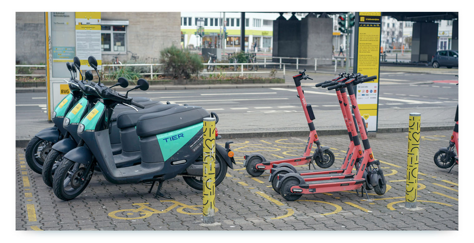 A public transportation station with multiple shared mobility options parked together. In the foreground, several gray and black Tier-branded electric scooters are parked next to a group of pink Voi-branded scooters. The area is marked with yellow bike and scooter symbols on the ground and yellow bollards. The image shows a designated mobility hub where different shared services are consolidated.
