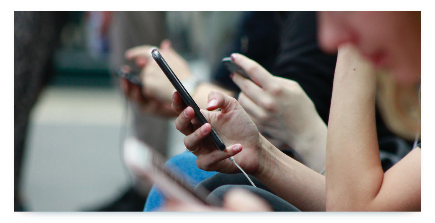 A close-up of a group of people holding smartphones. The central focus is on a person's hands holding a black smartphone with a white charging cable connected. The people are sitting, and their hands and devices are arranged closely, suggesting a crowd using their phones. This image represents the customer-facing technology aspect of a shared mobility service, such as the mobile app used for booking and managing rides.