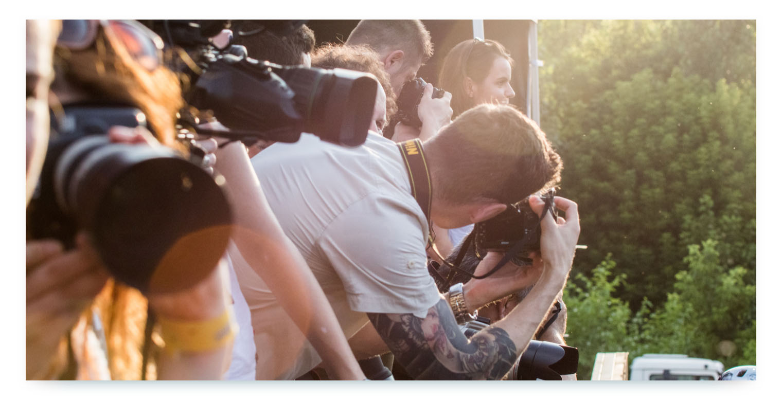 A close-up, low-angle shot of a crowd of photographers and journalists at an outdoor event. They are all holding large cameras with telephoto lenses, pointing them toward a subject that is not visible in the frame. The late afternoon sun shines brightly from the top right, creating a lens flare and casting a golden glow. The image represents the public relations aspect of a shared mobility service launch, with media attention and press coverage.