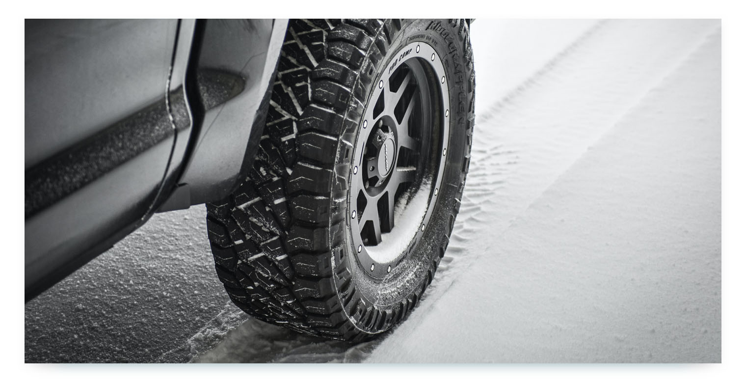 A close-up, black-and-white image of a car wheel with a tire designed for snowy conditions. The tire has a deep tread pattern and is visibly kicking up snow, while the car is driving on a partially snow-covered road. This image represents the seasonal maintenance of shared cars, such as changing tires to ensure safety and operational readiness in all weather conditions.