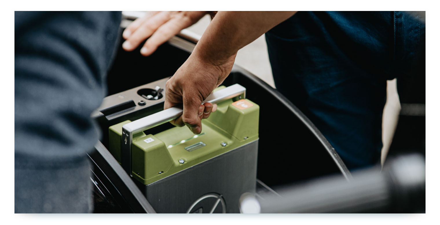 A close-up shot of two people's hands. One person is shown lifting a green and black electric vehicle battery out of a compartment in a car. This image illustrates the recurring process of swapping batteries to keep shared electric vehicles charged and operational.