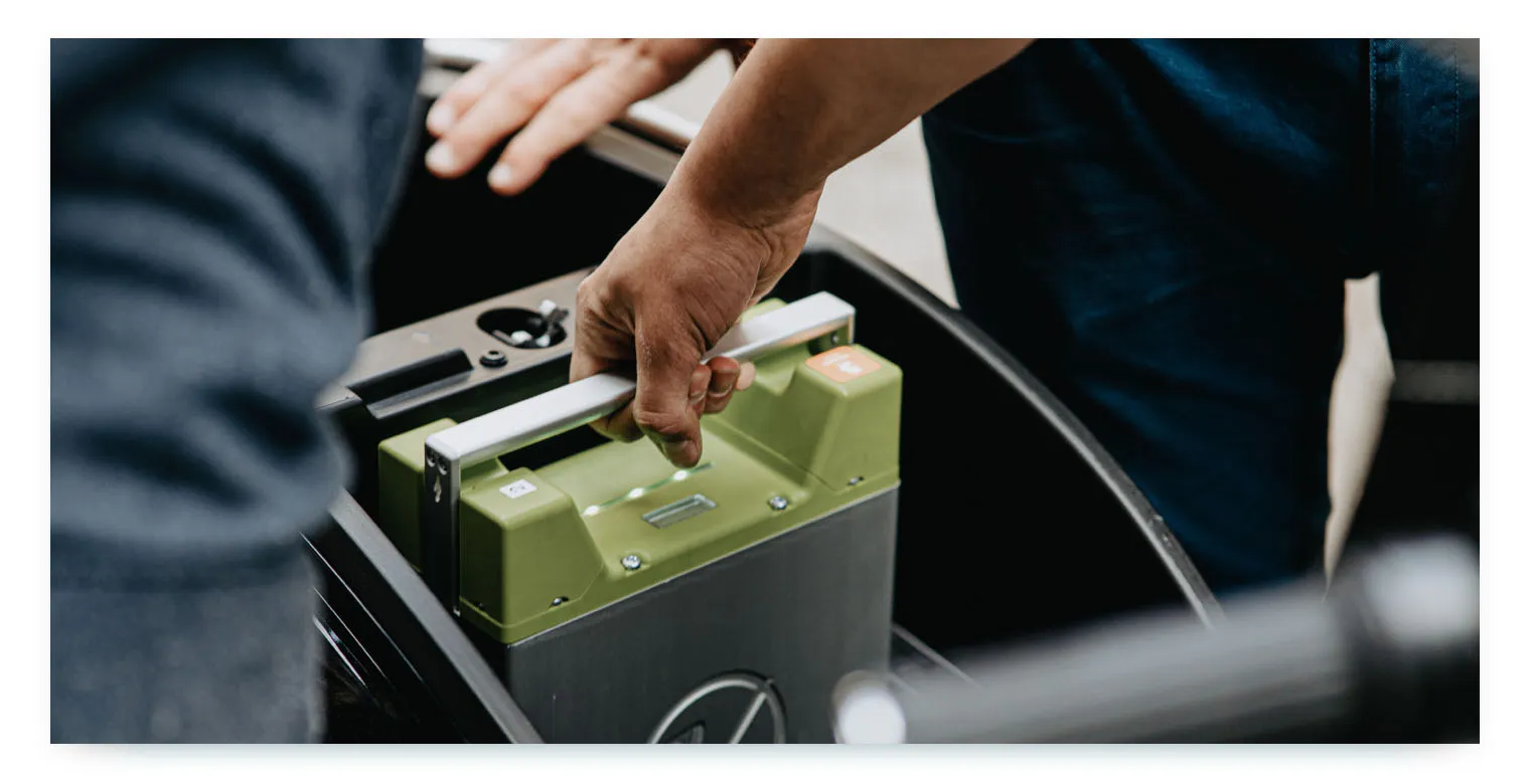 A close-up shot of two people's hands. One person is shown lifting a green and black electric vehicle battery out of a compartment in a car. This image illustrates the recurring process of swapping batteries to keep shared electric vehicles charged and operational.