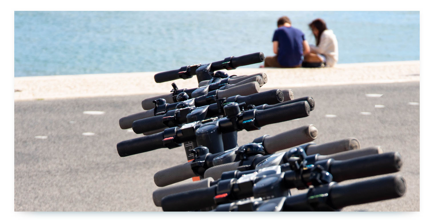 A close-up, low-angle shot showing the handlebars of a row of electric scooters parked in a line. The handlebars are in focus, while in the blurry background, a body of water and two people sitting on the ledge are visible. The image represents a fleet of shared e-scooters ready for use by customers, highlighting the need for efficient maintenance.