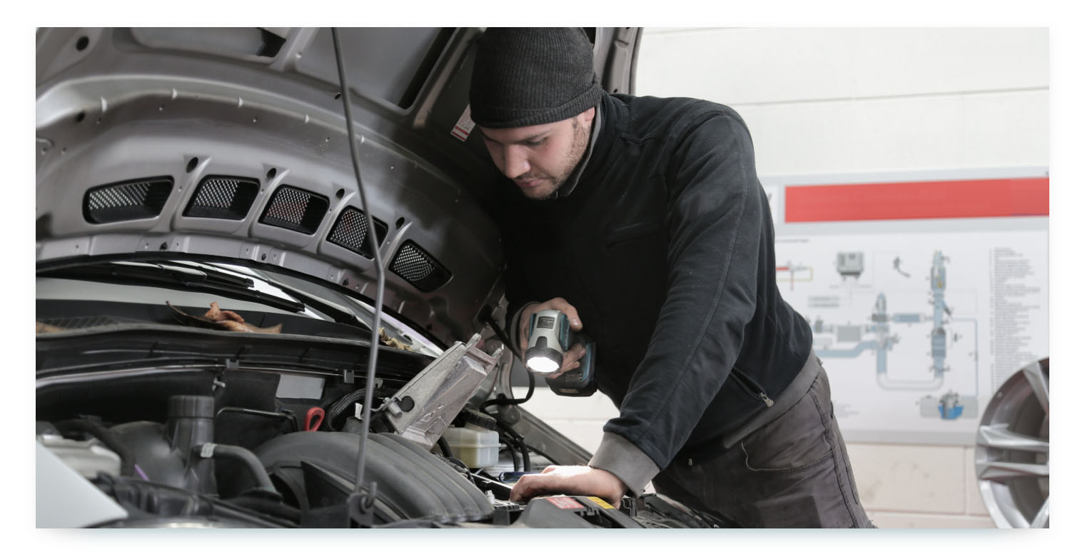 A professional mechanic, wearing a black beanie and black sweater, is shown from a side angle, leaning over the engine of a car. He holds a flashlight in one hand, illuminating a specific area of the engine as he inspects it. This image represents the important recurring process of vehicle inspection and maintenance to ensure the fleet's readiness and reliability.