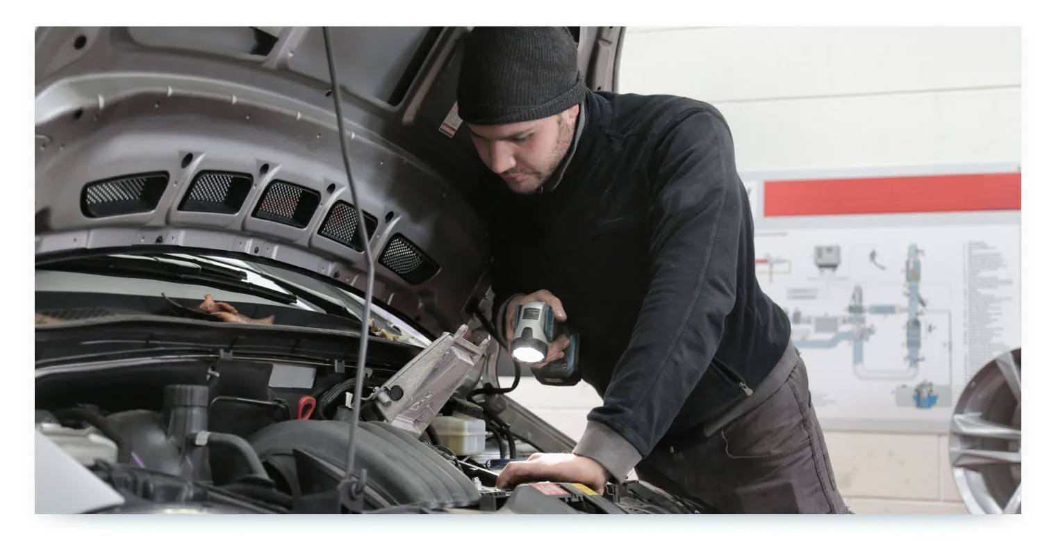 A professional mechanic, wearing a black beanie and black sweater, is shown from a side angle, leaning over the engine of a car. He holds a flashlight in one hand, illuminating a specific area of the engine as he inspects it. This image represents the important recurring process of vehicle inspection and maintenance to ensure the fleet's readiness and reliability.