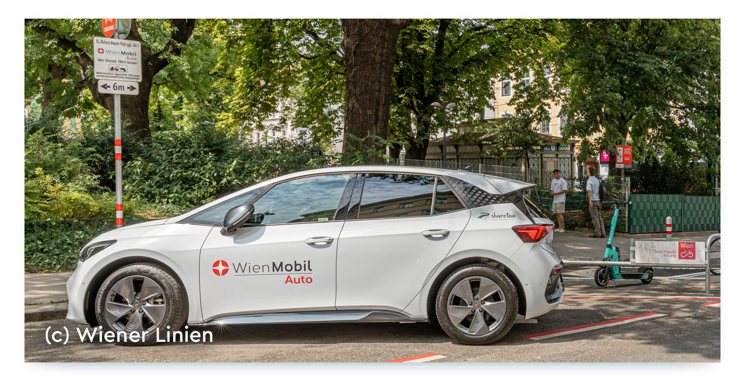 A white car from the carsharing service, with "WienMobil Auto" and the "sharetoo" logo on its side. It is parked on a city street in Vienna next to a sign and other micromobility vehicles like an electric scooter. The photo is credited to Wiener Linien.