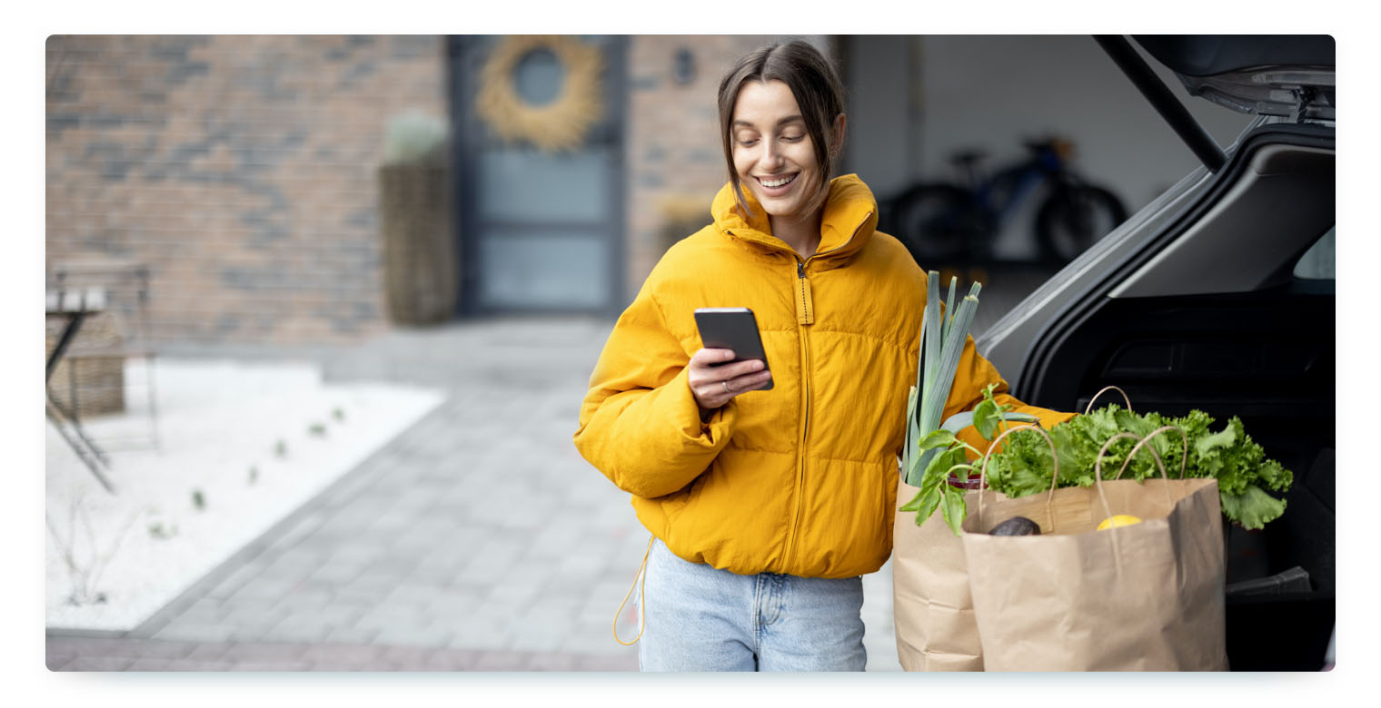 A woman wearing a yellow puffer jacket and jeans stands next to the open trunk of a car, which contains two paper grocery bags. She is holding a smartphone and smiling as she looks at the screen. The scene suggests the convenience of using a carsharing vehicle for errands.