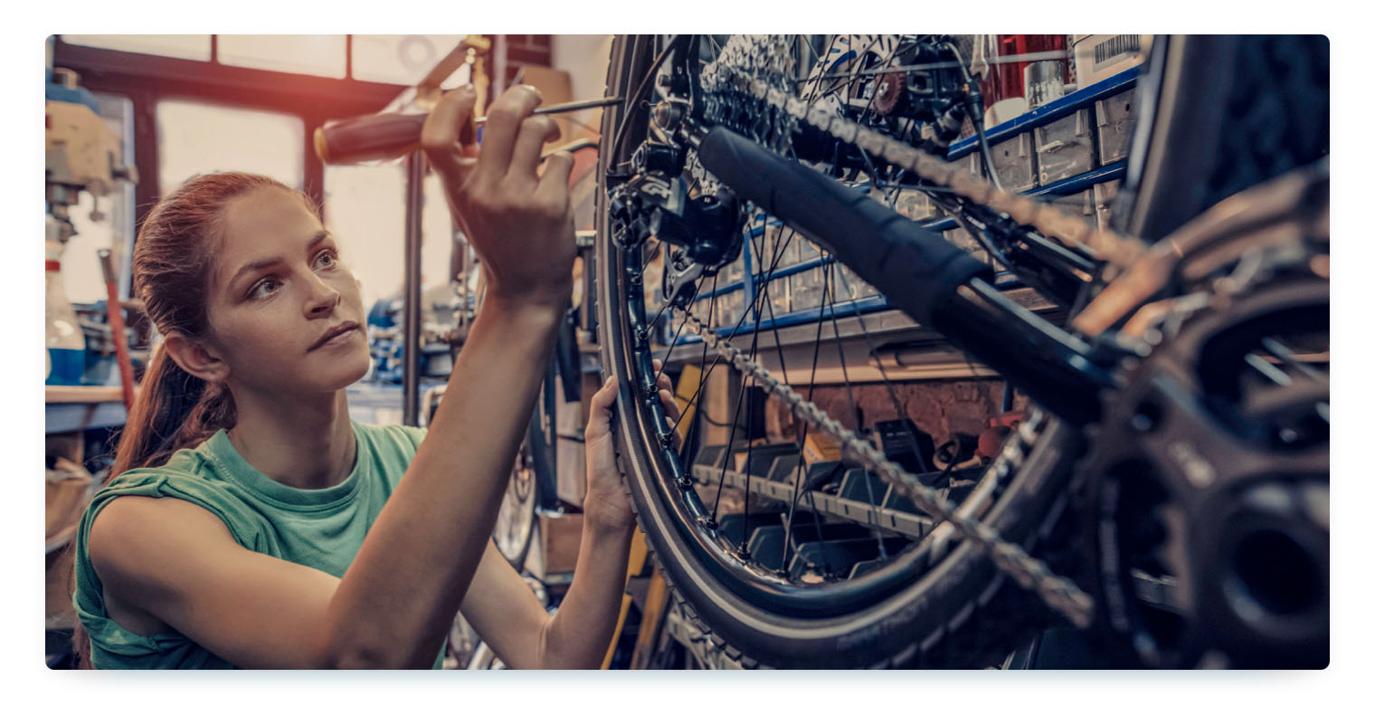 A woman is working on a bicycle in a workshop, focusing intently on adjusting the gears with a screwdriver. The photo is taken from a side angle and highlights the detailed work involved in vehicle maintenance and repair.