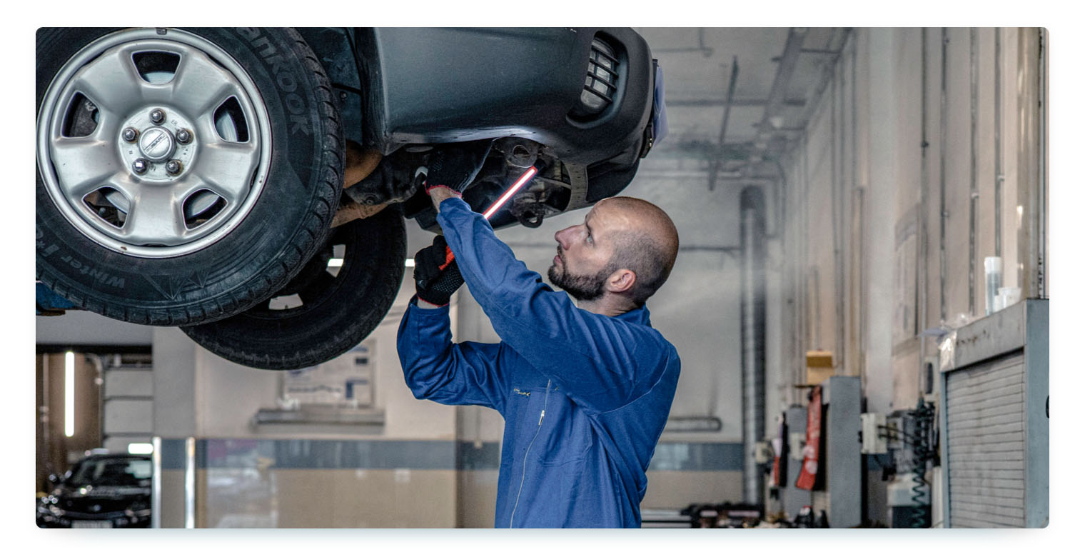 A mechanic in a blue jumpsuit is working under a car that is raised on a lift in a garage. He is holding a tool and looking up at the car's undercarriage, indicating a thorough vehicle inspection or repair process.